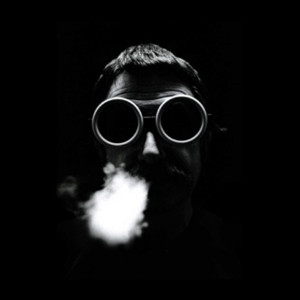 black and white photo of designer Sottsass blowing smoke out of mouth and wearing circle shared eye goggles